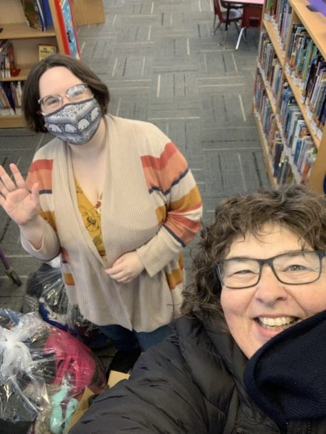 One woman in a mask smiling and waving at the camera while another woman takes a selfie. 